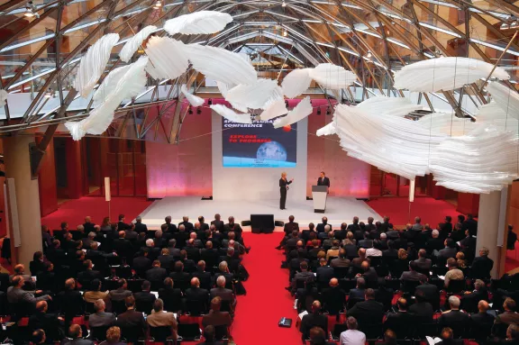 Veranstaltungsraum mit weißen Deckendekoration, roter Teppich und zwei Gruppen mit Sitzreihen, Bühne am Ende des roten Teppichs mit Präsentation