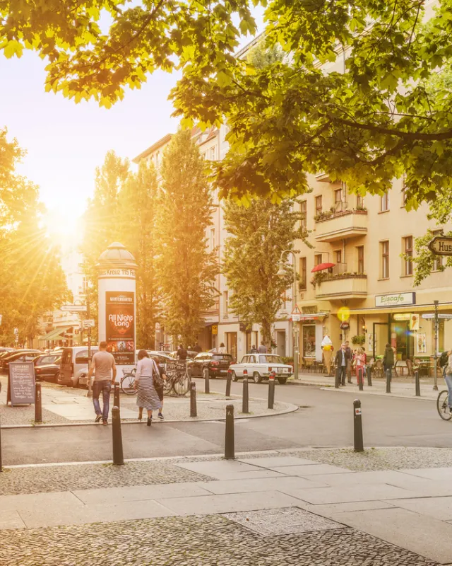 Kollwitzplatz bei Sonnenaufgang, einige PSaziergänger und Fahrradfahrer, grüne Bäume und 