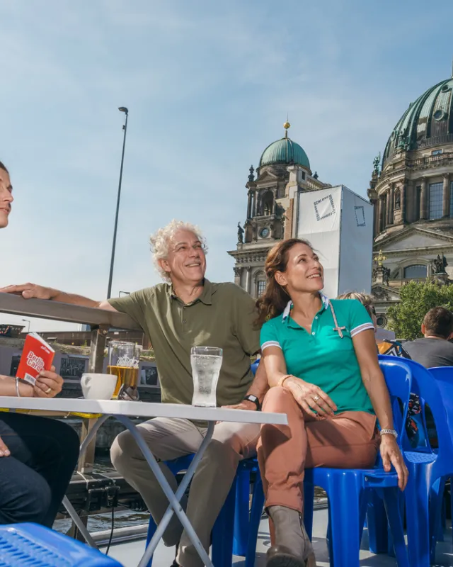 Links sitzende Frau mit blauem Hemd und Berlin WelcomeCard in der Hand an Tisch, rechts Frau mit grünem Shirt und Mann mit braunem Oberteil sitzen auch an Tisch, blaue Plastikstühle rechts im Bild, Berliner Dom im Hintergrund, alle schauen nach rechts oben 