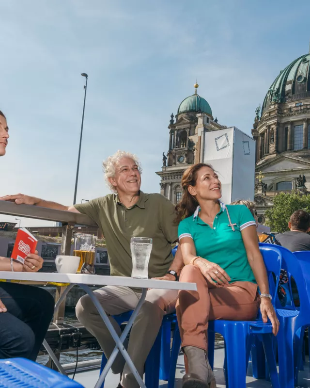 Links sitzende Frau mit blauem Hemd und Berlin WelcomeCard in der Hand an Tisch, rechts Frau mit grünem Shirt und Mann mit braunem Oberteil sitzen auch an Tisch, blaue Plastikstühle rechts im Bild, Berliner Dom im Hintergrund, alle schauen nach rechts oben 