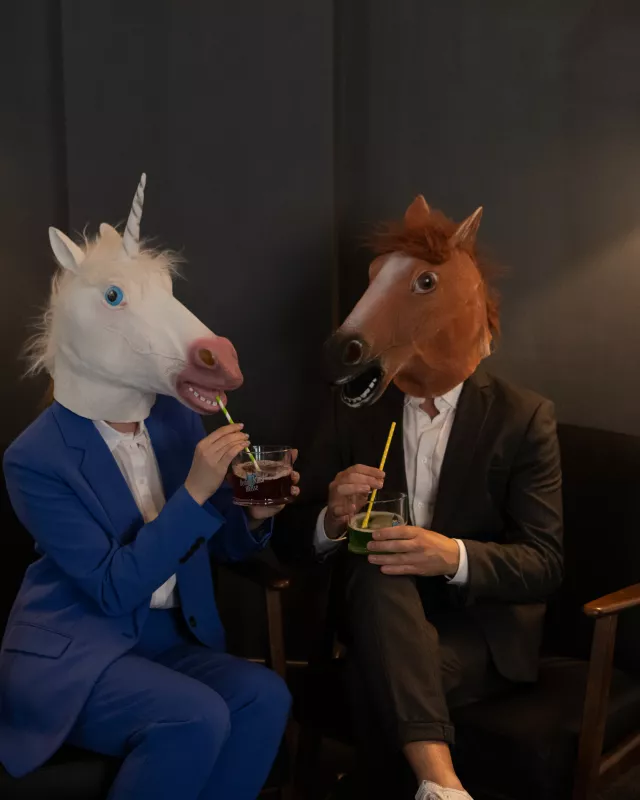 Zwei Menschen in Anzügen mit Pferdemasken auf dem Kopf und Drink in der Hand