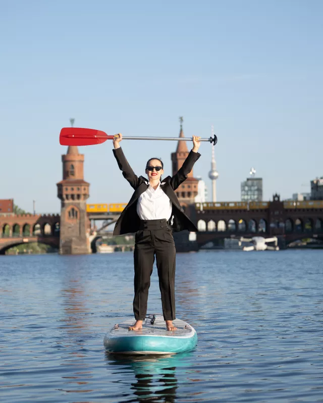 Frau in Business Anzug auf SUP auf der Spree die das Paddel mit beiden armen in die Höhe hält