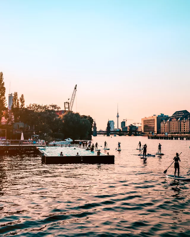 People Paddleboarding In Spree River By City Against Clear Sky During Sunset