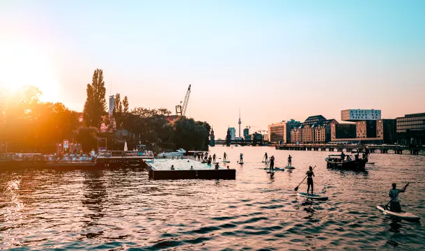 People Paddleboarding In Spree River By City Against Clear Sky During Sunset
