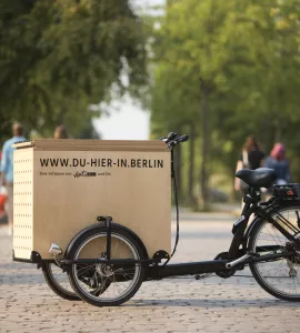 schwarzes Lasten-Fahrrad mit großer Holzbox auf Weg, Bäume säumen Weg