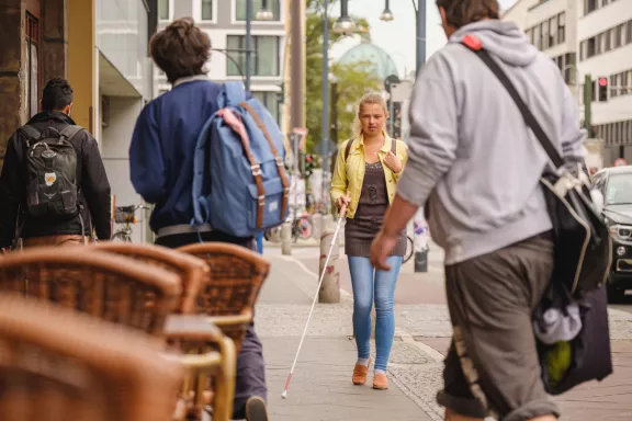 Berlin-Barrierefrei Frau mit Blindenstock läuft auf Gehsteig Kamera entgegen, drei weitere Menschen mit Rücken zu Kamera,