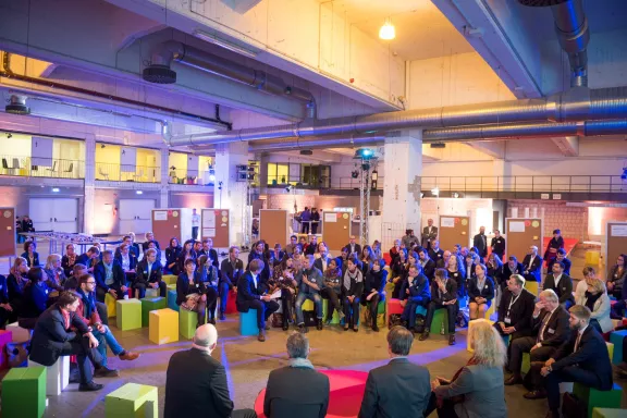Berlin MICE Summit 2016 Maschinenhalle, viele Menschen sitzen im lockeren Stuhlkreis auf bunten Hockern, bläulich gedimmtes Licht