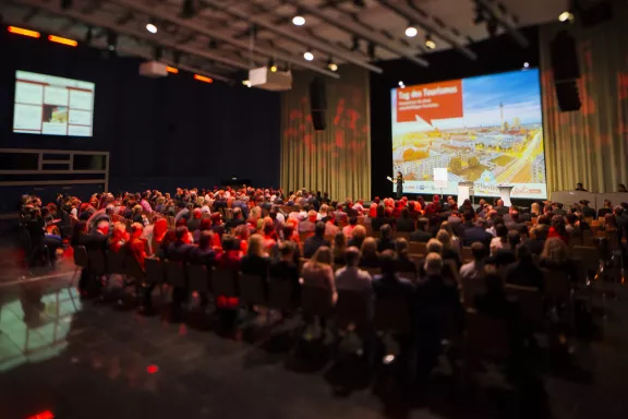 Tag des Tourismus 2015, Menschen in Reihenbestuhlung, Blick auf Leinwand & Bühne mit visitBerlin Präsentation, dunkles Licht