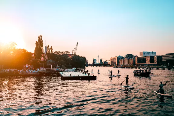 People Paddleboarding In Spree River By City Against Clear Sky During Sunset